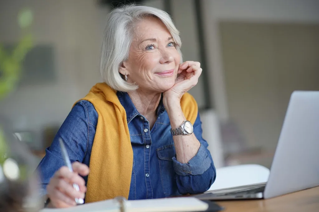 A mature adult women with white hair, dressed in business casual attire, sitting in front of a laptop computer at a desk in a modern office environment, following along and engaging in a work meeting with business colleagues.